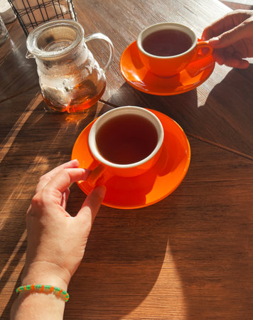 Hands are holding bright orange tea cups on a wooden table, accompanied by a glass teapot, illuminated by warm sunlight, creating a cozy and inviting sceneの写真素材