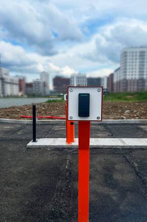 Urban scene with a black device for security parking system on an orange pole, set against a blurred city skyline and overcast sky, highlighting modern design and infrastructureの写真素材
