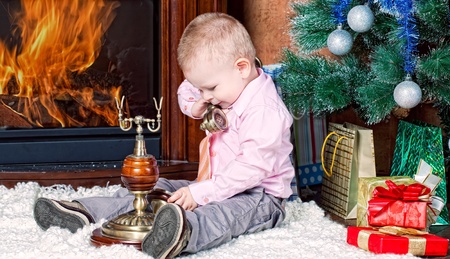little boy in a room with a fireplace sits at a Christmas fur-tree and calls by phoneの写真素材
