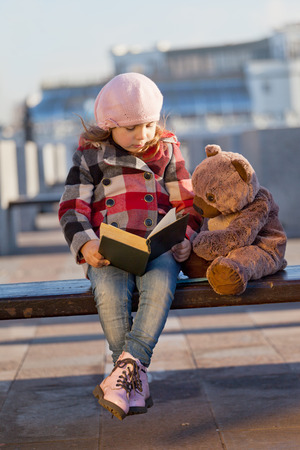 Girl sits on a bench and reads the book to a toy bearの写真素材