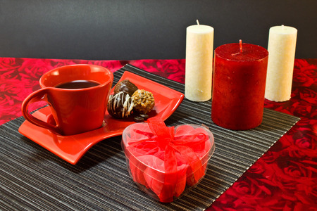 Coffee in red cup on a saucer with a chocolate, a present in a box in the form of heart on the background of candlesの写真素材
