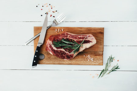 Raw ribeye steak on board, knife, fork, pepper, rosemary and pink Himalayan salt
