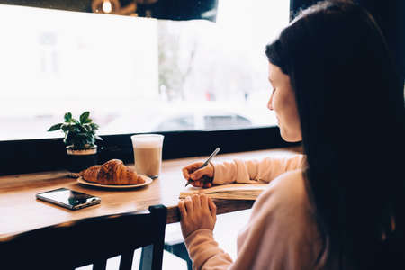 Girl writing in notebook, croissant, coffee, phone on wooden table. Blurredの写真素材