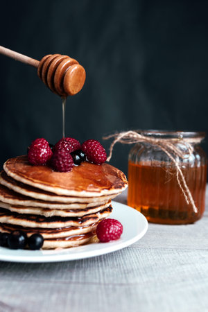Juicy pancakes with berries and honey on a white plate, spoon, jar, wooden table. High quality photo