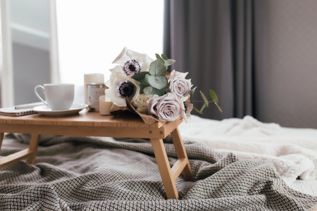 Romantic morning. Wooden coffee table with flowers on bed with plaid, coffee cup, flowers and candles. Lilac roses with eucalyptus and anemones. Interior gray tones. High quality photoの写真素材