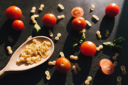 Red cherry tomatoes, green basil, butterfly vermicelli pasta, wooden spoon on gray background. High...