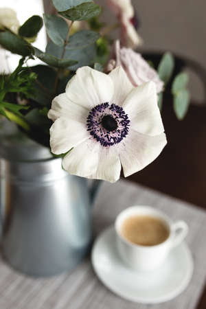 Delicate anemones flowers in a metal pot vase with a cup of coffee on the wooden table. Blurred backgroundの写真素材