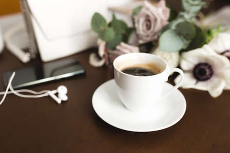 A cup of coffee with a bouquet of tender anemone, eucalyptus branches and pastel purple roses, a woman handbag, headphones and a phone on a brown wooden table. Blurred backgroundの写真素材