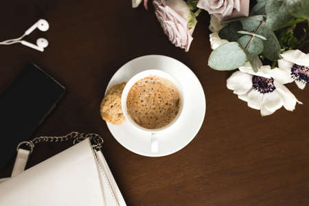 A cup of coffee with a bouquet of tender anemone, eucalyptus branches and pastel purple roses, a woman handbag, headphones and a phone on a brown wooden table. Blurred backgroundの写真素材