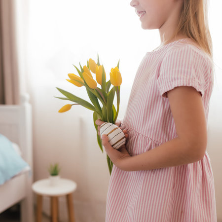 Easter concept. Small smiling girl in white fluffy bunny ears and pink dress holding yellow tulip...