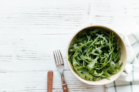Green fresh arugula salad leaves in bowl, fork and knife on white wooden background, white towel....