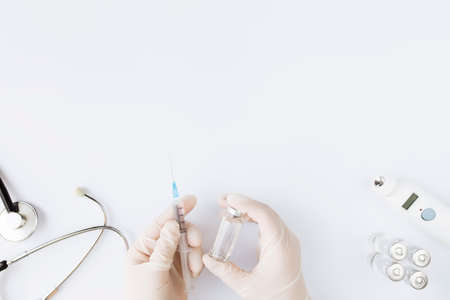 Medicine treatment, vaccination concept. Doctor hands holding bottle with vaccine and injection syringe, electronical thermometer, stethoscope and vaccine vial on white background. High quality photoの写真素材