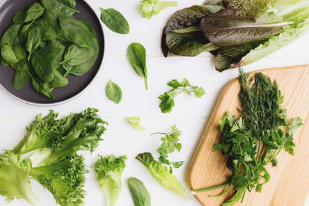 Flat lay with cutting board and bowl of fresh green salad leaves of spinach and lettuce, romaine and parsley, basil on white background. Healthy vegetarian eating concept. High quality photoの写真素材