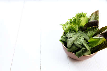Flat lay with bowl of fresh green salad leaves of spinach and lettuce, romaine and basil on white wooden background. Healthy vegetarian eating concept. High quality photoの写真素材