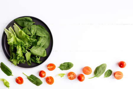 Flat lay with bowl of fresh green salad leaves, lettuce, spinach and red cherry tomatoes cups and halves on a white background. Healthy vegetarian eating concept. High quality photoの写真素材