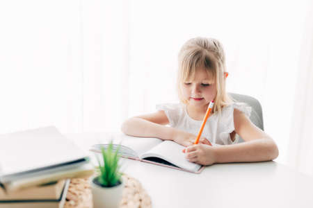 A little girl sits at the table and writes in a white notebook. Education concept. Home schooling. Homework. High quality photoの写真素材