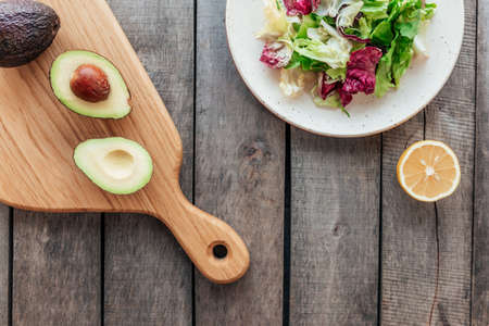 Healthy eating concept flat lay. Mediterranean diet, plate with fresh green lettuce salad leaves, purple radicchio leaves, cutting board with avocado halves, kernel, lemon on wooden backgroundの写真素材