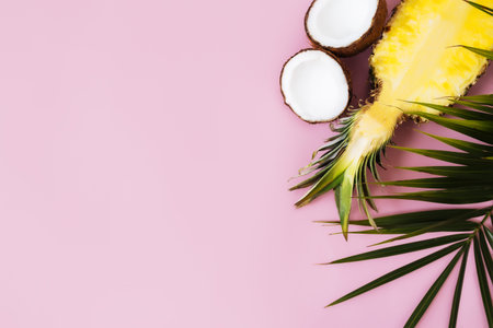 Flat lay with cut halves of fresh pineapple, coconut and green palm leaf on a pastel pink...