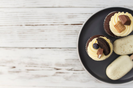 White cookie ice creams on a stick and tasty muffins and cupcakes on a tray on a wooden background. Decorated with different candies, cookies and colored sweet cream cheese on top.の写真素材