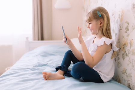 A little girl sits on the bed in the stylish bedroom, holding phone and have a video call using smartphone, waving a hand. Communication or homeschooling conceptの写真素材