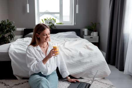 Pretty smiling Caucasian girl sits on the floor in bedroom and use laptop for remote work and drink orange juice. Communication, technology, remote work concepts in gray interior apartmentの写真素材