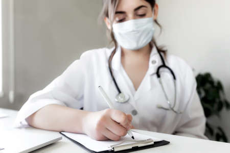 The doctor in shite dress with stethoscope and face protective mask writes something in the patient's medical history. High quality photoの写真素材