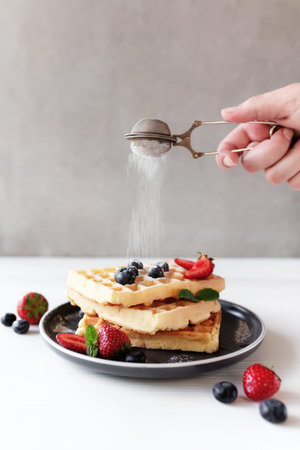 Hand is shaking powdered sugar over stack of waffles on a plate on the table with blueberry, chopped...