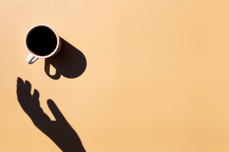 White cup and saucer plate of black espresso coffee from top view with shadow of sun and hand holding or touching the cup on yellow beige terracotta background. High quality photoの写真素材