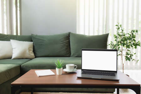 Work from home, workspace, desktop, remote work concept, gray thin laptop computer on brown wooden table with white cup of coffee, green sofa, flowerpot, note pad. Stylish apartments comfort zoneの写真素材