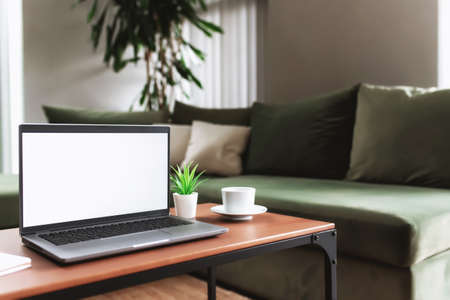 Work from home, workspace, desktop, remote work concept, gray laptop computer with empty white screen mock up on brown wooden table with white cup of coffee, note pad, flowerpot, blurred backgroundの写真素材
