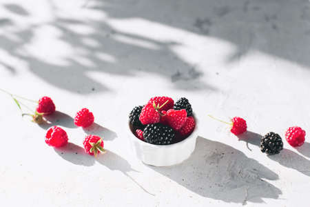 White bowl full with raspberries, blackberry on gray concrete table with leaves, tree branches shadow. Healthy eating concept. Eco, bio farming. Fresh tasty berries on gray background. Quality photoの写真素材