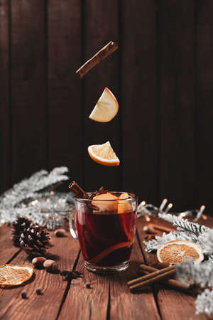 Christmas glass cup or mug with Mulled Wine on wooden table and background with levitating cinnamon stick, dried or sublimated orange, apple slices, anise, spices, hazelnuts, decoration light garlandsの写真素材