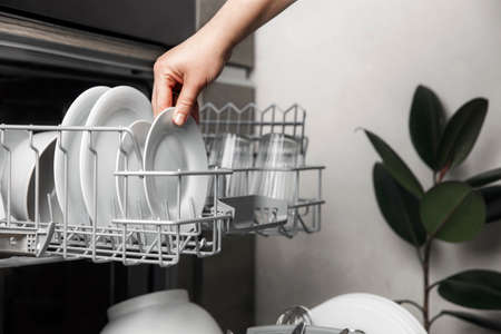 Close-up of female hand loading dished to, empty out or unloading from open automatic stainless built-in dishwasher machine with clean utensils inside in modern home kitchen. Household domestic lifeの写真素材