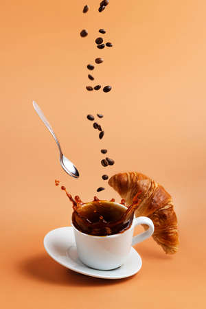 White coffee cup, plate and metal tea spoon falling on beige table background and coffee splashes because of hit, coffee beans levitate, fly and fall to cup, crispy fresh croissant. Breakfast conceptの写真素材
