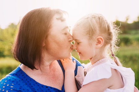 Grandmother holding granddaughter on her arms and kissing nose. Happy childhood, family love concept. Support and togetherness. Two people outdoor with sunlight background. High quality photoの写真素材