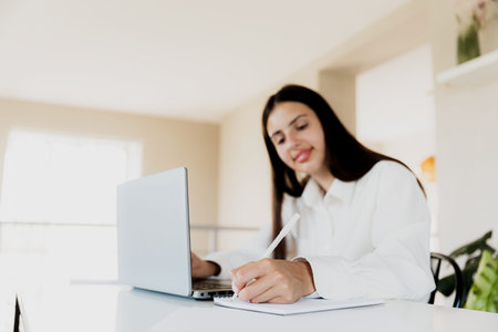 Caucasian business woman sitting at desk, reading from computer, taking notes at notebook. Female...