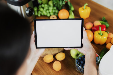 Female hands holding tablet with empty blank white screen mockup in front of vegetables, fruits on table in kitchen at home. Selective focus, cropped. Food delivery concept, mobile diet plan appの写真素材