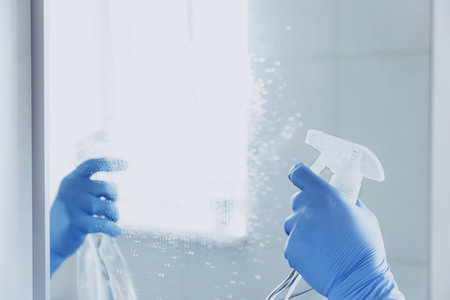 Cropped view of housekeeper woman hand in rubber blue gloves spraying chemical cleaner liquid to the mirror while cleaning bathroom at home. Housekeeping and cleaning service. Clean house, cleanlinessの写真素材
