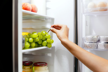 Woman hand taking, grabbing or picks up green bunch of grapes out of open refrigerator shelf or fridge drawer full of fruits, blueberries, bottles with yogurt. Healthy food diet, lifestyle conceptの写真素材