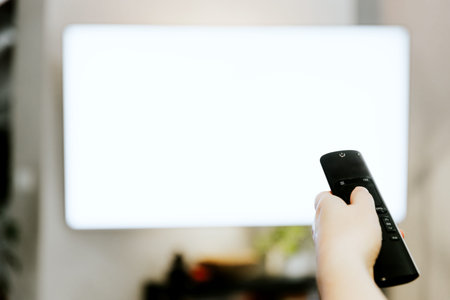 Hand holding remote control pointing to TV. Person watching smart television indoors. White empty...
