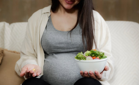 Close up of young pregnant woman holding bowl of salad and pill in hand, receiving vitamins, taking dietary supplements at home. Pregnancy, prenatal health and wellness, healthy nutrition, eatingの写真素材