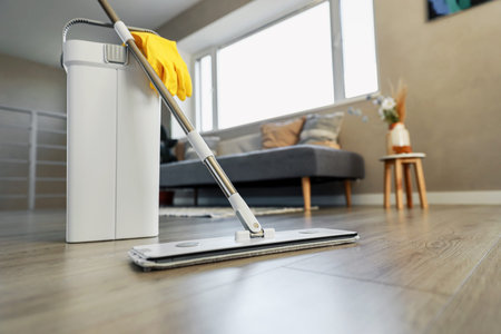 Cleaning supplies: mop, gloves, bucket with wringer sitting on wooden laminate floor in modern...
