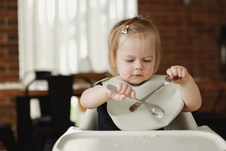 Eleven month infant girl or toddler sitting on high chair at feeding table. Kid holding spoon in hand. Baby child learning to eat by itself. Making a mess. Solid introduction, development conceptの写真素材