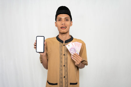 Asian Muslim man wearing brown Muslim shirt smiling happily while holding money and showing...