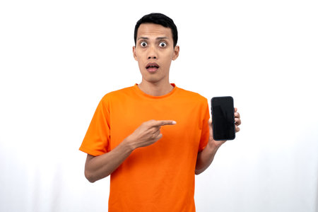 Portrait of a young Asian man wearing an orange t-shirt with a happy, surprised, pleased expression while pointing at a smartphone screen. Isolated on a white background.の写真素材