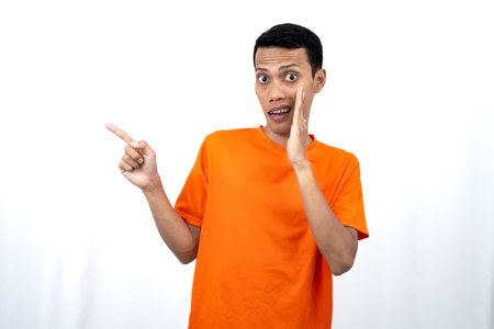 Portrait of a young Asian man wearing an orange t-shirt with his hand near his mouth in a whispering pose while pointing to the side of the copy space. Isolated on white background.の写真素材