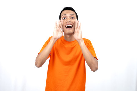 Portrait of a young Asian man wearing an orange t-shirt with his hand near his mouth while sharing news isolated on white background.の写真素材