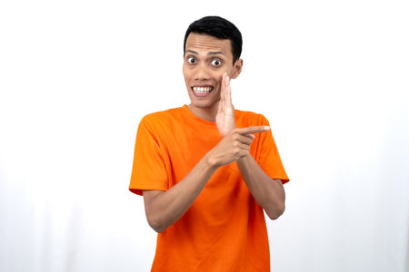 Portrait of a young Asian man wearing an orange t-shirt with his hand near his mouth in a whispering pose while pointing to the side of the copy space. Isolated on white background.の写真素材