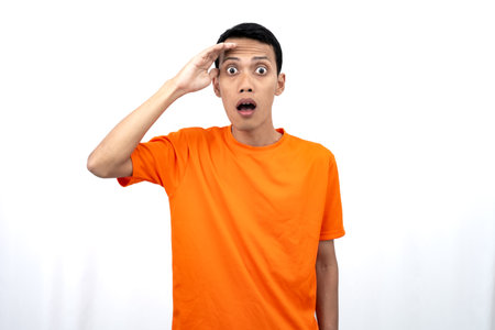 Portrait of a young Asian man wearing an orange t-shirt looking surprised, shocked and shocked. Hands are near eyes, isolated on white background.の写真素材