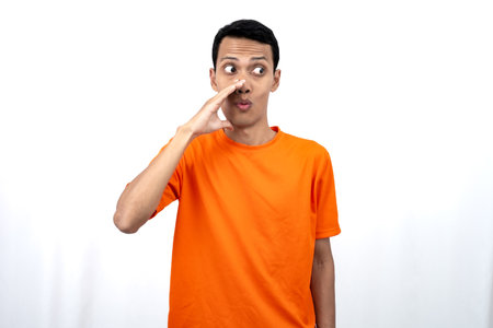 Portrait of a young Asian man wearing an orange t-shirt with his hand near his mouth whispering gossip and telling secrets while sharing news isolated on white background.の写真素材